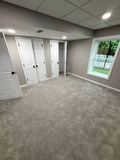 A finished basement room with gray carpet, double closet doors, a window and recessed lights in a drop ceiling