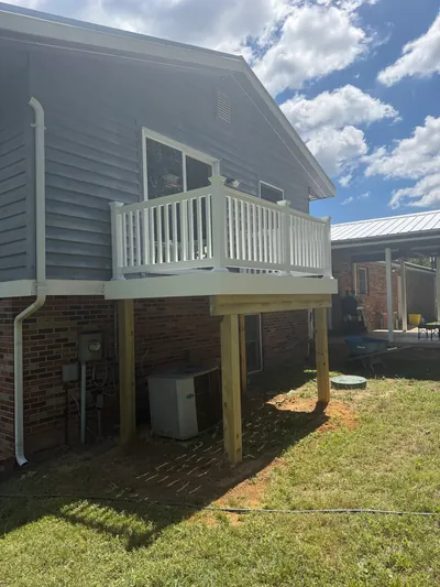 A raised deck with white railing on square wood posts at the back of a gray-sided house with a brick lower level