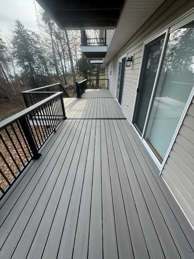A long run of composite decking with black railing along the back of a house, below a balcony