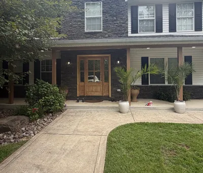 The front of a two-story house with stacked stone on its gable and entry, a wood front door and a porch on square posts