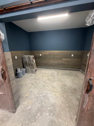 The back room of the garage addition seen through open barn doors: navy walls over wood plank wainscot, a strip light and a bare concrete floor with paint cans and flooring boxes