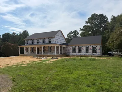 A two-story home under construction, wrapped and roofed, with a long front porch on white columns and a garage wing to the right