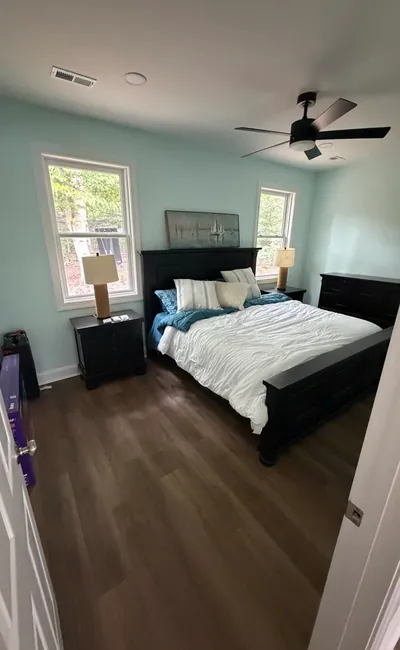 A bedroom in the pool house with pale blue walls, two windows, a ceiling fan and wood-look flooring
