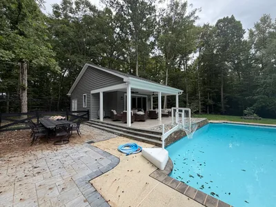 A gray pool house with a covered patio on white posts, set at the end of a swimming pool with a paver deck