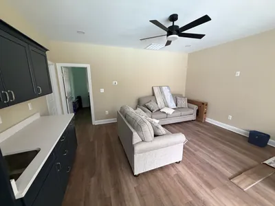 Inside the pool house: a sitting room with two sofas and a black ceiling fan, and a kitchenette counter with dark cabinets along the left wall