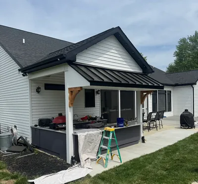 A covered porch with a black standing seam metal roof on wood brackets, over an outdoor counter with a grill
