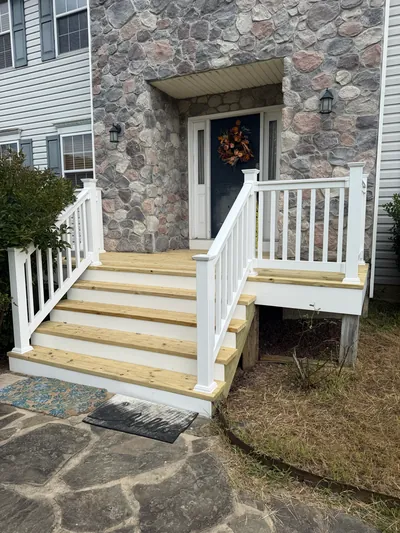 New wood steps and a landing with white railing, at a front door set in a stone wall