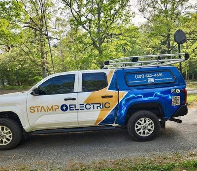 A white pickup with a blue cap and a gold lightning bolt wrap that reads Stamp Electric, parked on gravel under green trees
