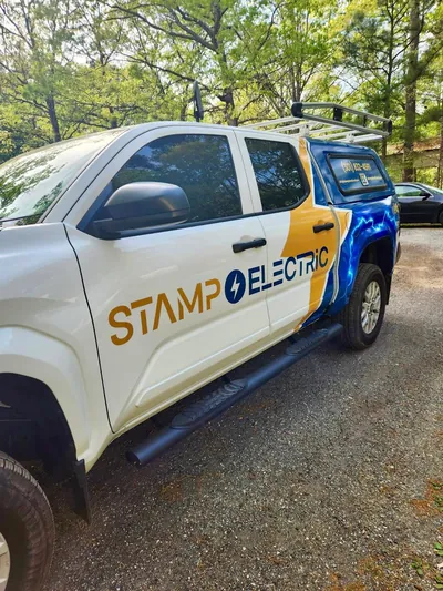 The Stamp Electric truck from the front: a white pickup with a gold and blue lightning bolt wrap and a ladder rack, parked on gravel under trees